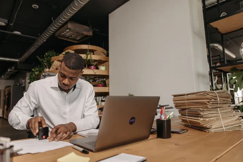 A man stamping documents at an office desk beside a laptop