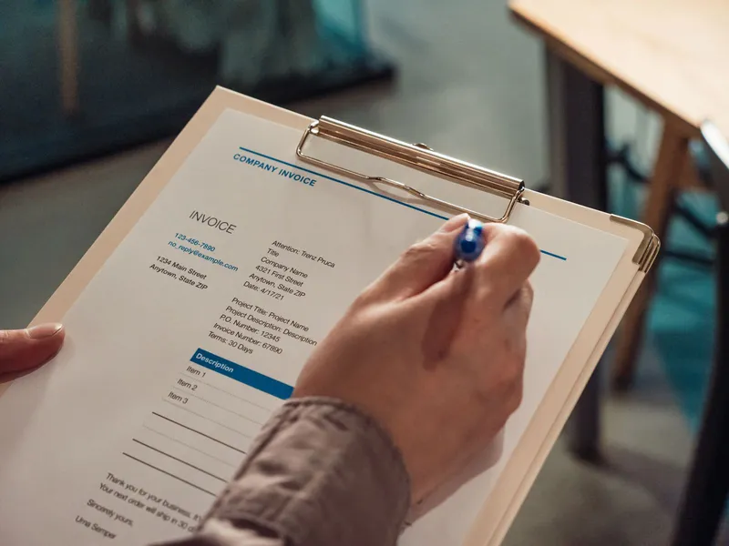 A hand holding an invoice on a clipboard with a pen